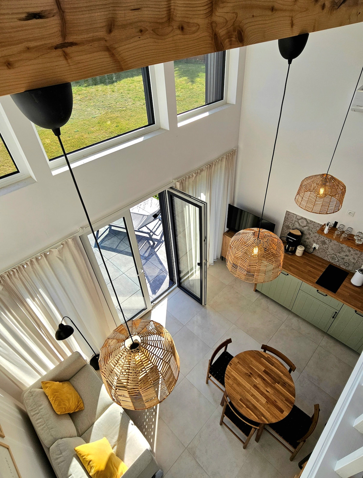 A modern living space features large windows allowing natural light to flood in. Rattan pendant lights hang from the ceiling above a wooden dining table and chairs, complemented by a cozy sofa adorned with yellow cushions. Sliding glass doors lead to an outdoor seating area.