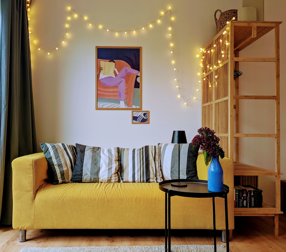 A bright yellow sofa with striped cushions is accompanied by a small black side table. A blue vase holds fresh flowers, while a painting hangs on the wall above. Soft string lights add a warm glow to the cozy seating area.