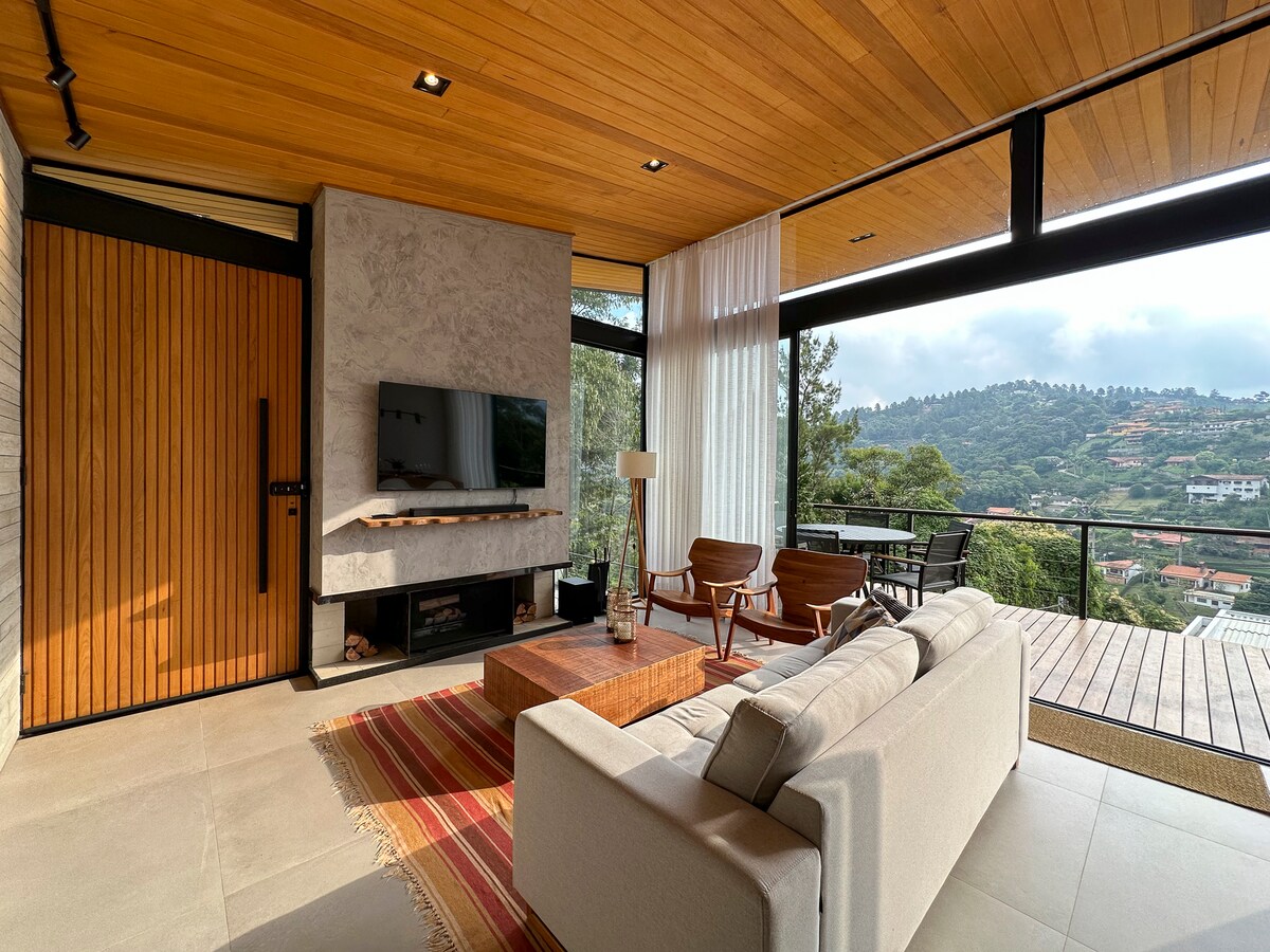 A modern living area showcases a soft, neutral sofa paired with mid-century style chairs. A wooden coffee table sits at the center, complementing the wood-paneled walls. Large windows provide an expansive view of the surrounding greenery and distant hills, with soft lighting enhancing the space.