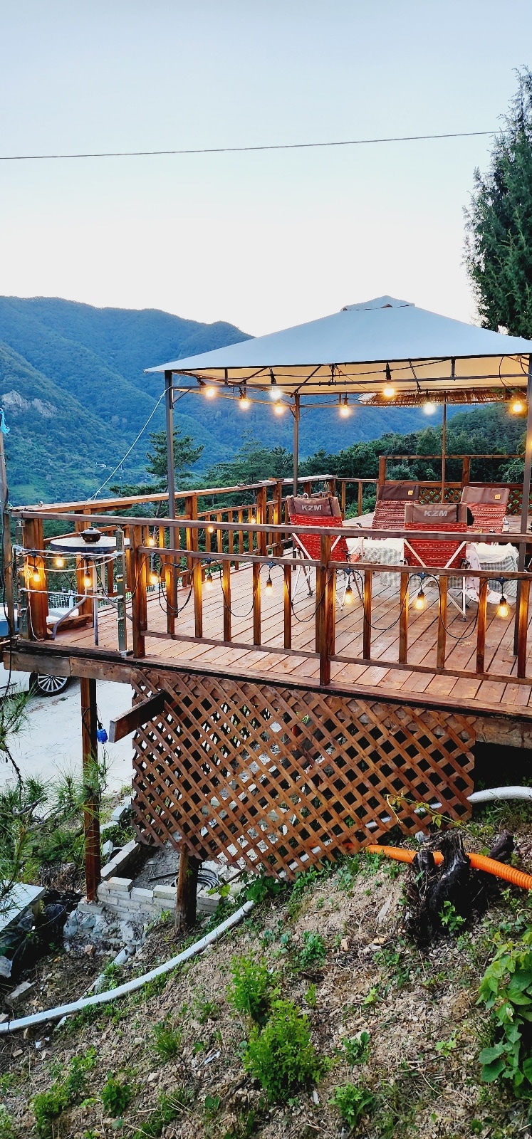 An outdoor deck is featured, furnished with two red chairs and a shaded gazebo adorned with string lights. An inviting atmosphere is created by the surrounding greenery and the scenic mountain backdrop, providing a tranquil space for relaxation.
