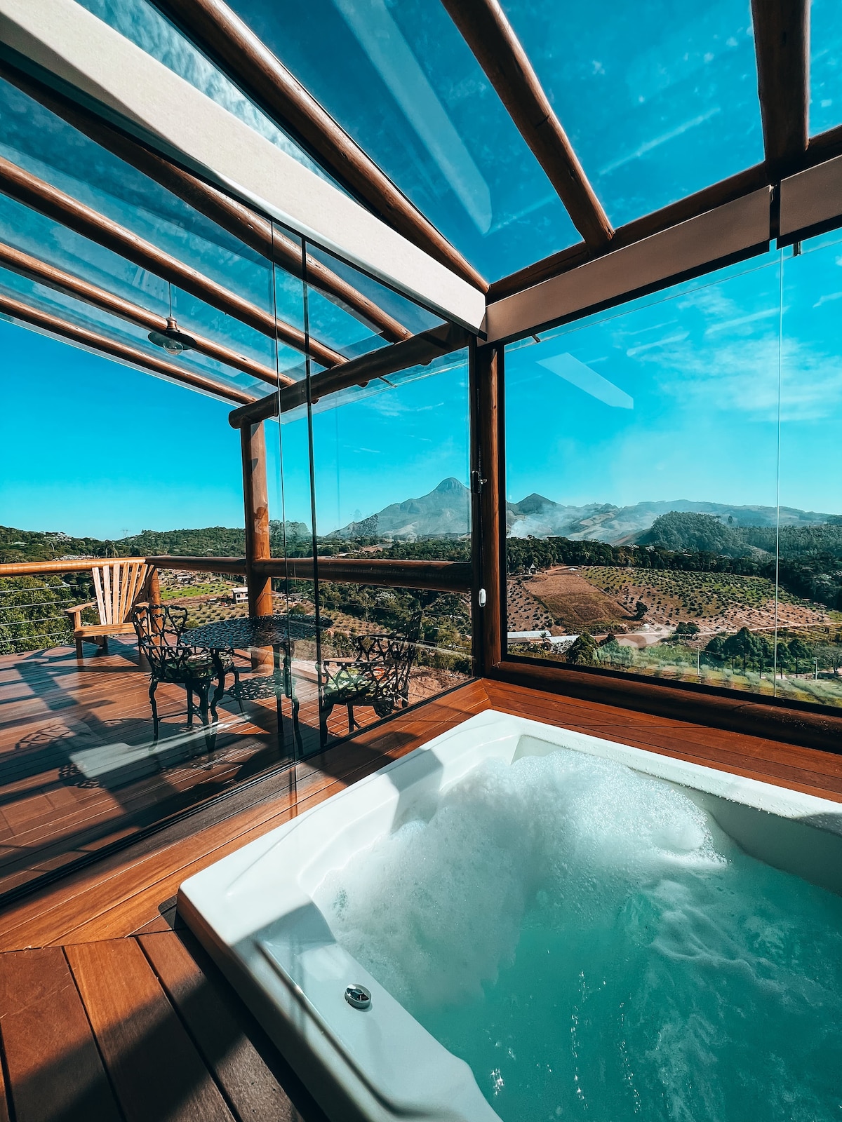A private hot tub is positioned beside large glass panels that provide a panoramic view of the surrounding mountains. An outdoor seating area with wooden chairs complements the space, highlighting the inviting blend of indoor and outdoor experiences.