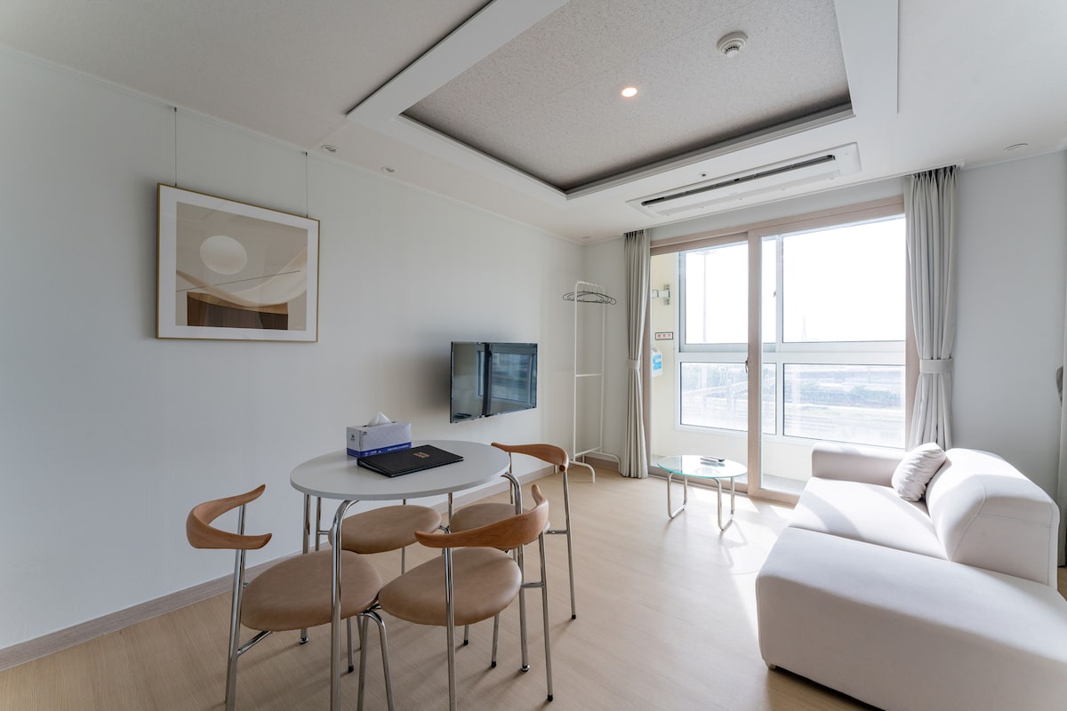 A bright living area is featured with ample natural light streaming through large windows. A sleek white sofa is positioned beside a round dining table with four wooden chairs. A wall-mounted TV is visible, enhancing the modern design of the space.