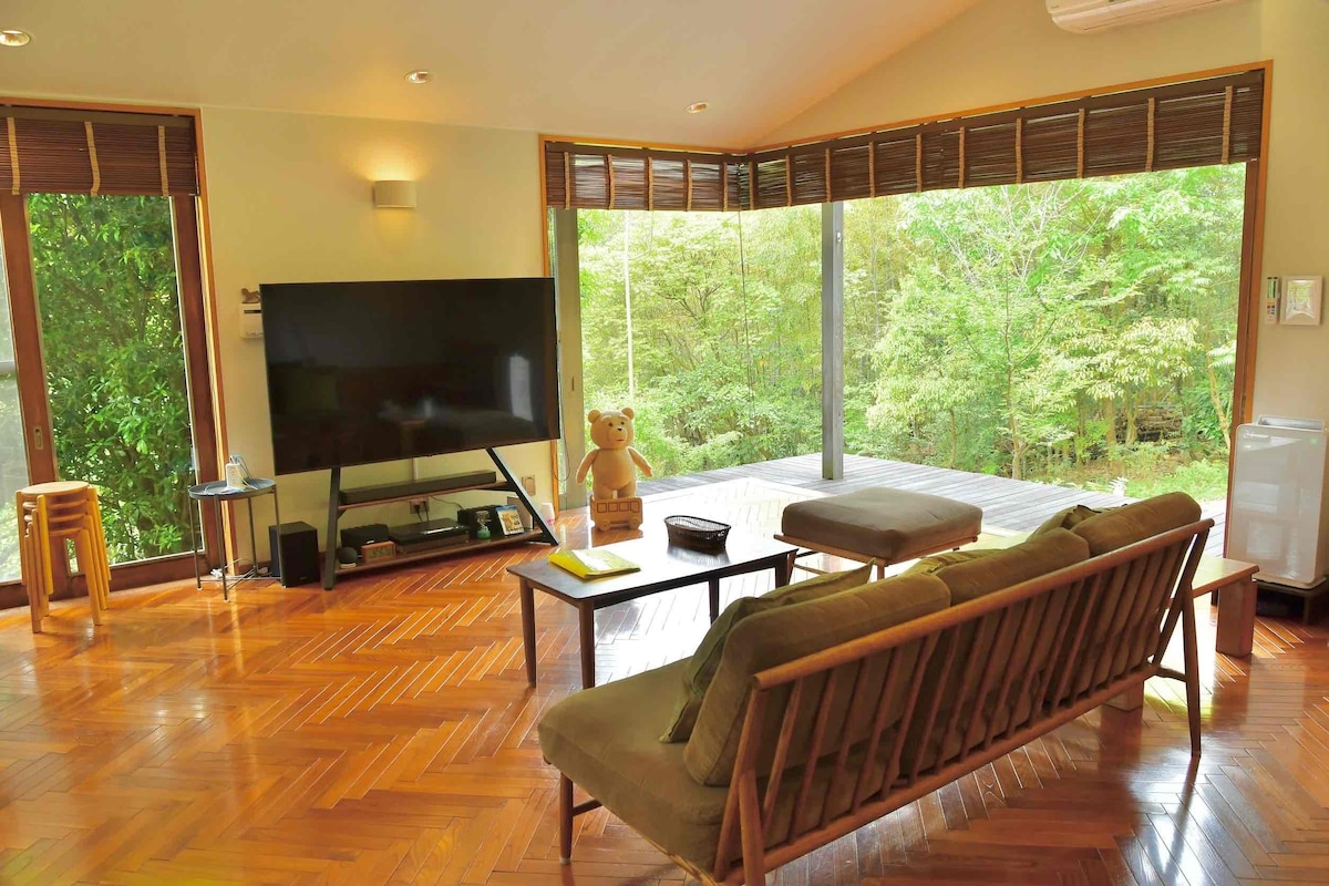 A spacious living area is depicted, featuring an inviting wooden sofa and a coffee table. Large glass doors offer a view of the lush greenery outside, enhancing natural light. A television stands in the corner, while a teddy bear adds a playful touch to the ambiance.