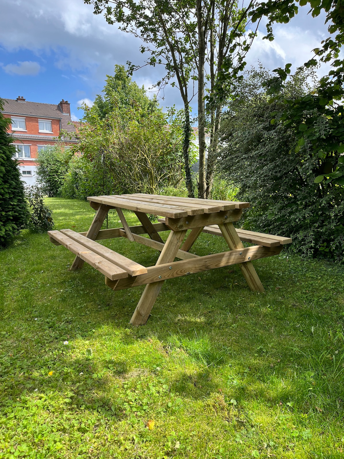 A wooden picnic table is placed on a grassy area, surrounded by greenery, including tall trees and shrubs. The setting offers a peaceful outdoor space ideal for relaxation or dining.