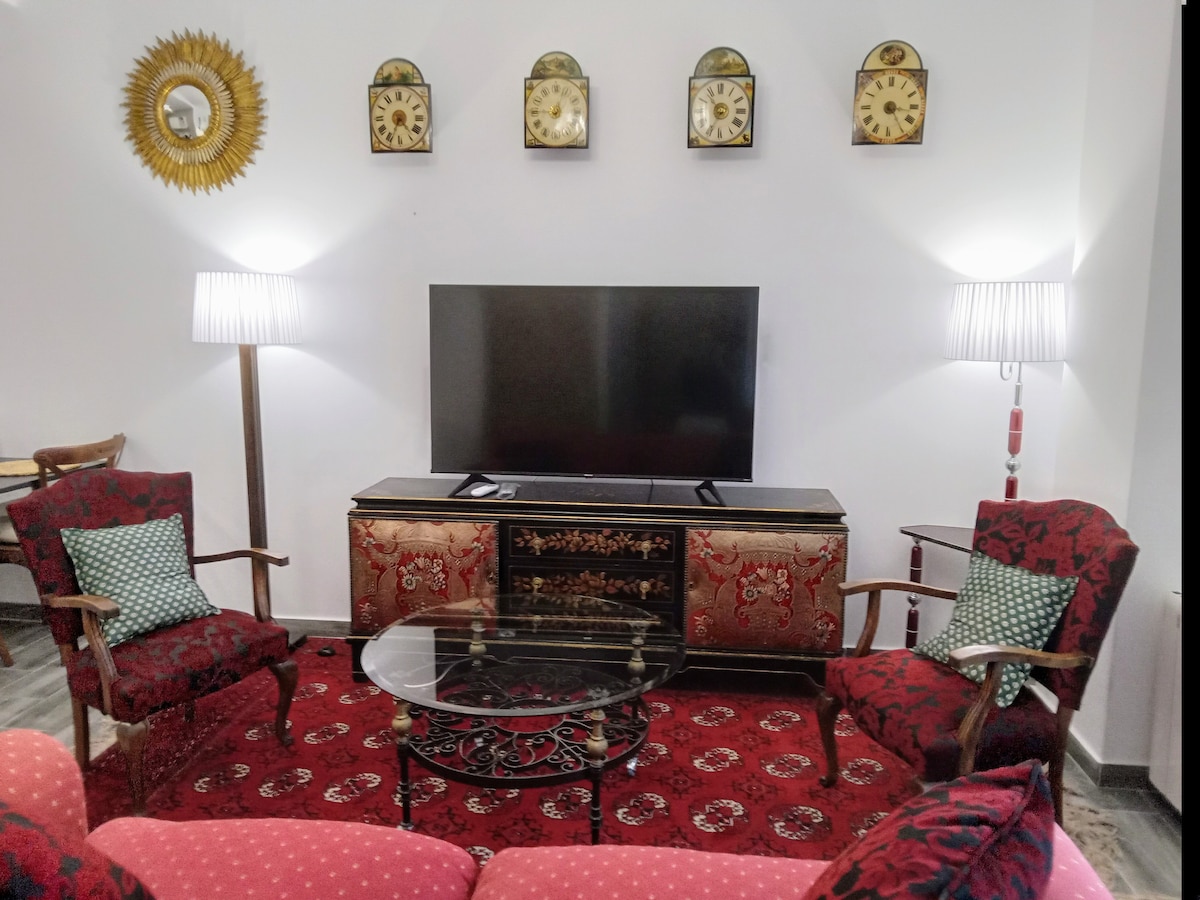A cozy seating area features two red patterned armchairs and a plush red sofa, arranged around a glass-top coffee table. A large television sits on an antique-style cabinet beneath decorative clocks, with soft light provided by two standing lamps.