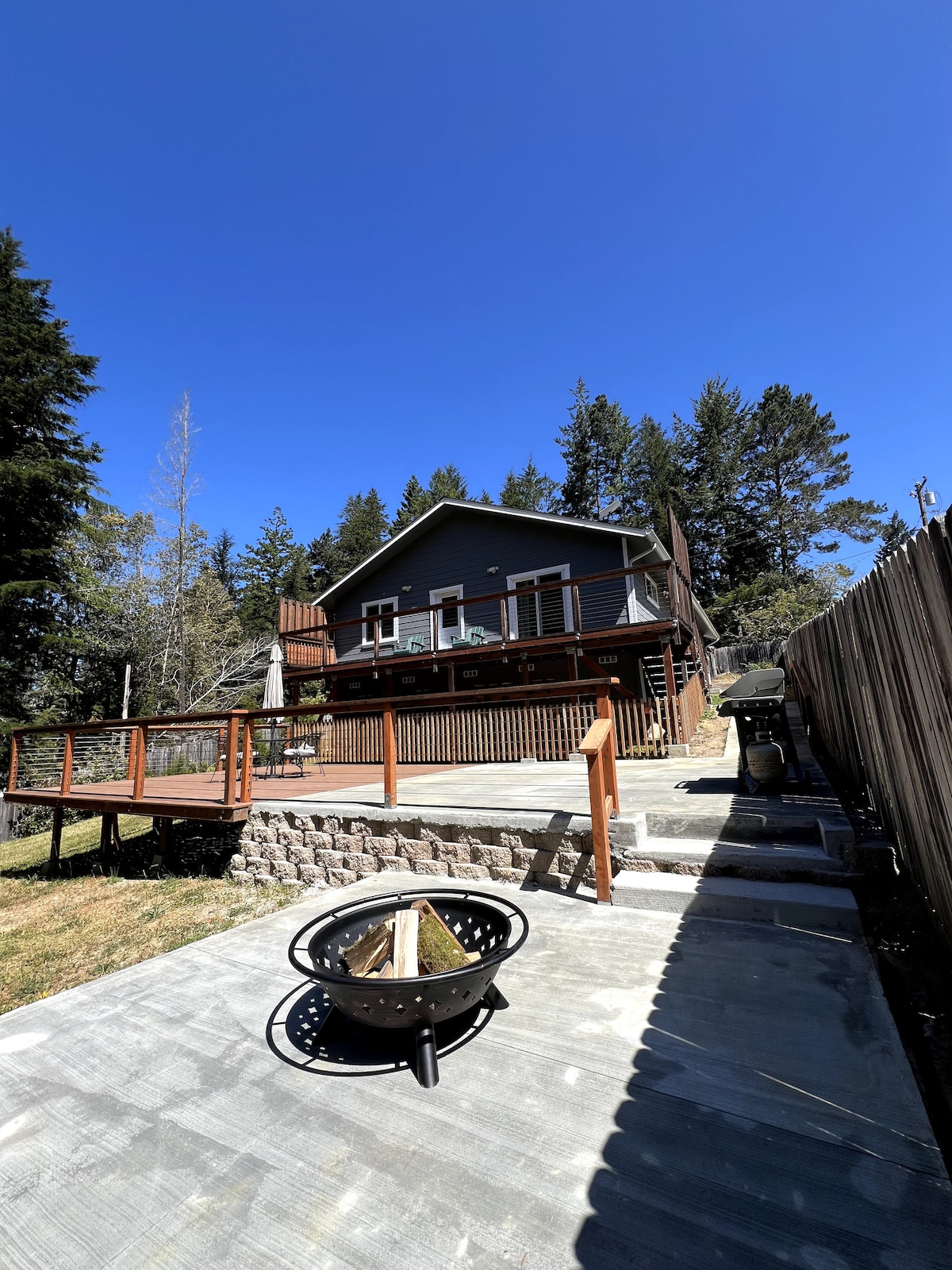 A two-story house is depicted, featuring a wooden deck with a railing and a patio umbrella. A fire pit with logs is placed on a stone patio, and natural greenery surrounds the area. The sky is clear with bright sunlight illuminating the scene.