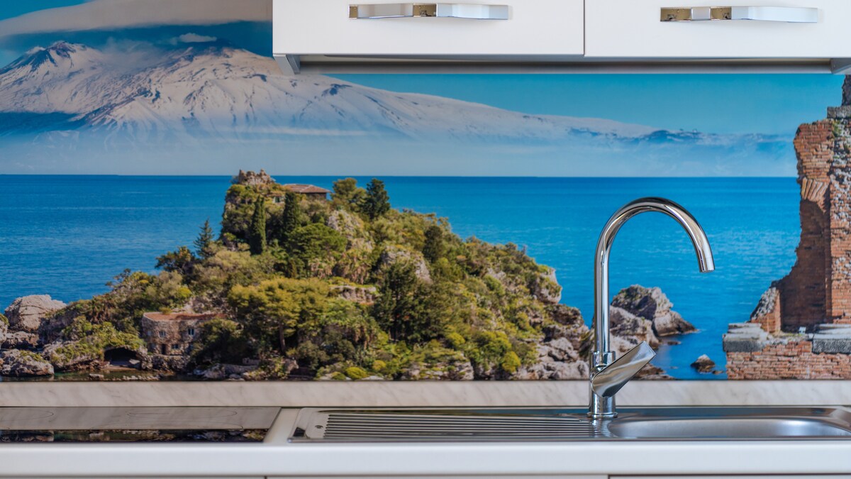 A modern kitchen sink is set against a vibrant backsplash depicting a coastal landscape featuring rocky shores and plants, with a view of the sea and distant mountains. Clean lines and a sleek faucet are visible, complementing the coastal theme.