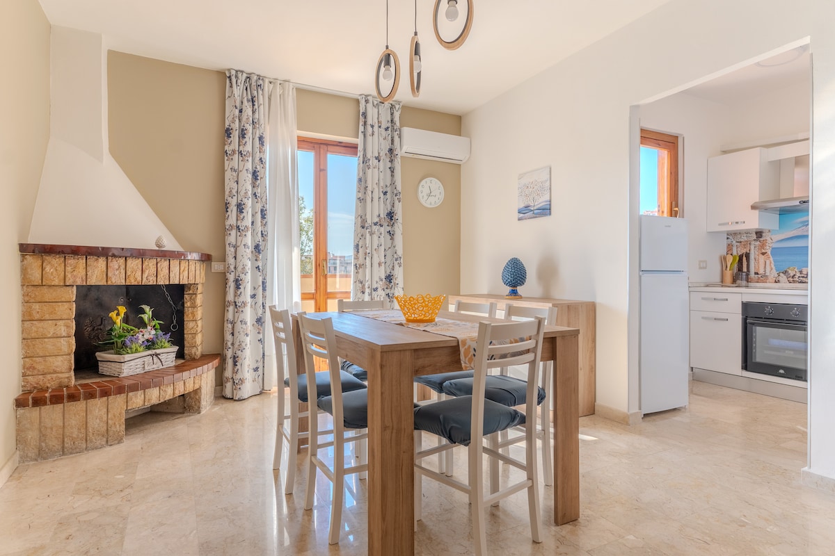 A spacious dining area features a wooden table surrounded by white chairs. A decorative fireplace is positioned against one wall, while a modern kitchen is visible in the background. Natural light streams in through the window, accentuating the light-colored walls.