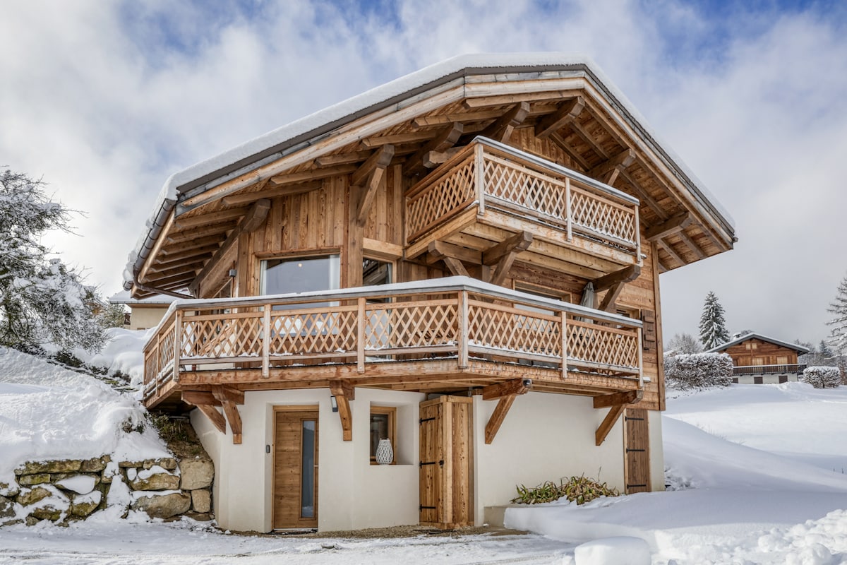 The exterior of the chalet showcases a traditional wooden design, featuring a sloped roof with overhanging eaves. Balconies with ornate wooden railings are visible on the upper levels, while snow blankets the surrounding area, enhancing the serene alpine atmosphere.