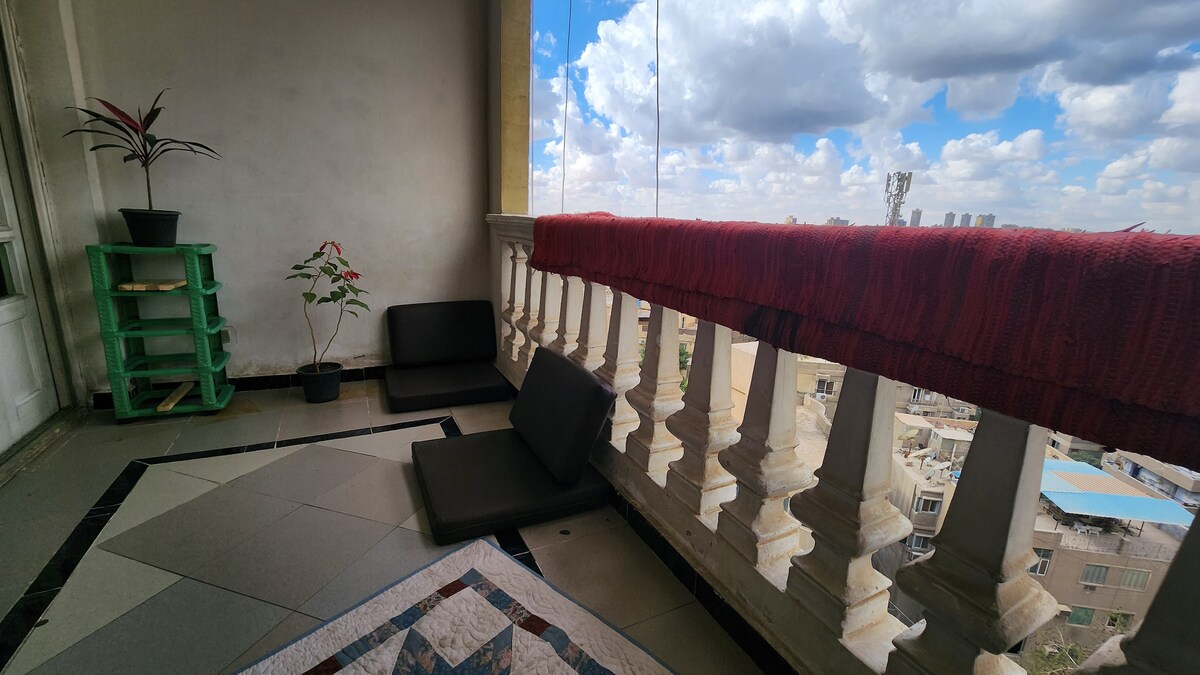 A balcony space features low seating cushions positioned along the railing, offering views of the city below. A small plant is displayed beside the seating area and a vibrant textile drapes over the railing. The floor is decorated with geometric tiles.