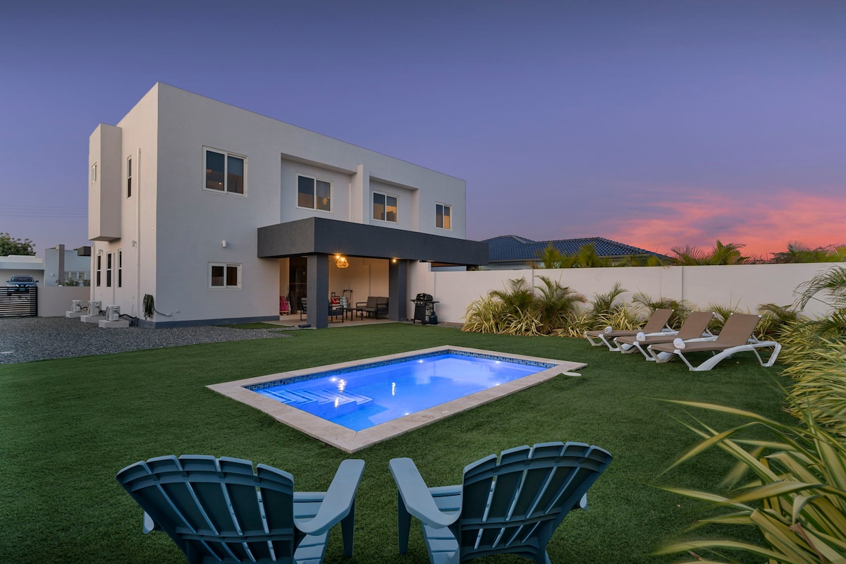 The exterior of the modern home is showcased against a colorful sunset sky. A private pool is surrounded by lush greenery and lounge chairs, while two light blue Adirondack chairs provide a cozy seating area near the pool.