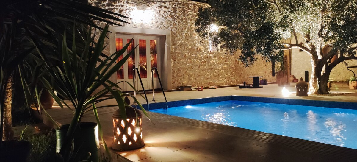 A serene swimming pool is surrounded by charming greenery and stone walls, illuminated softly by warm lighting. A decorative lantern is placed nearby, enhancing the relaxing atmosphere. The pool area features steps leading into the water, while trees create a peaceful ambiance.
