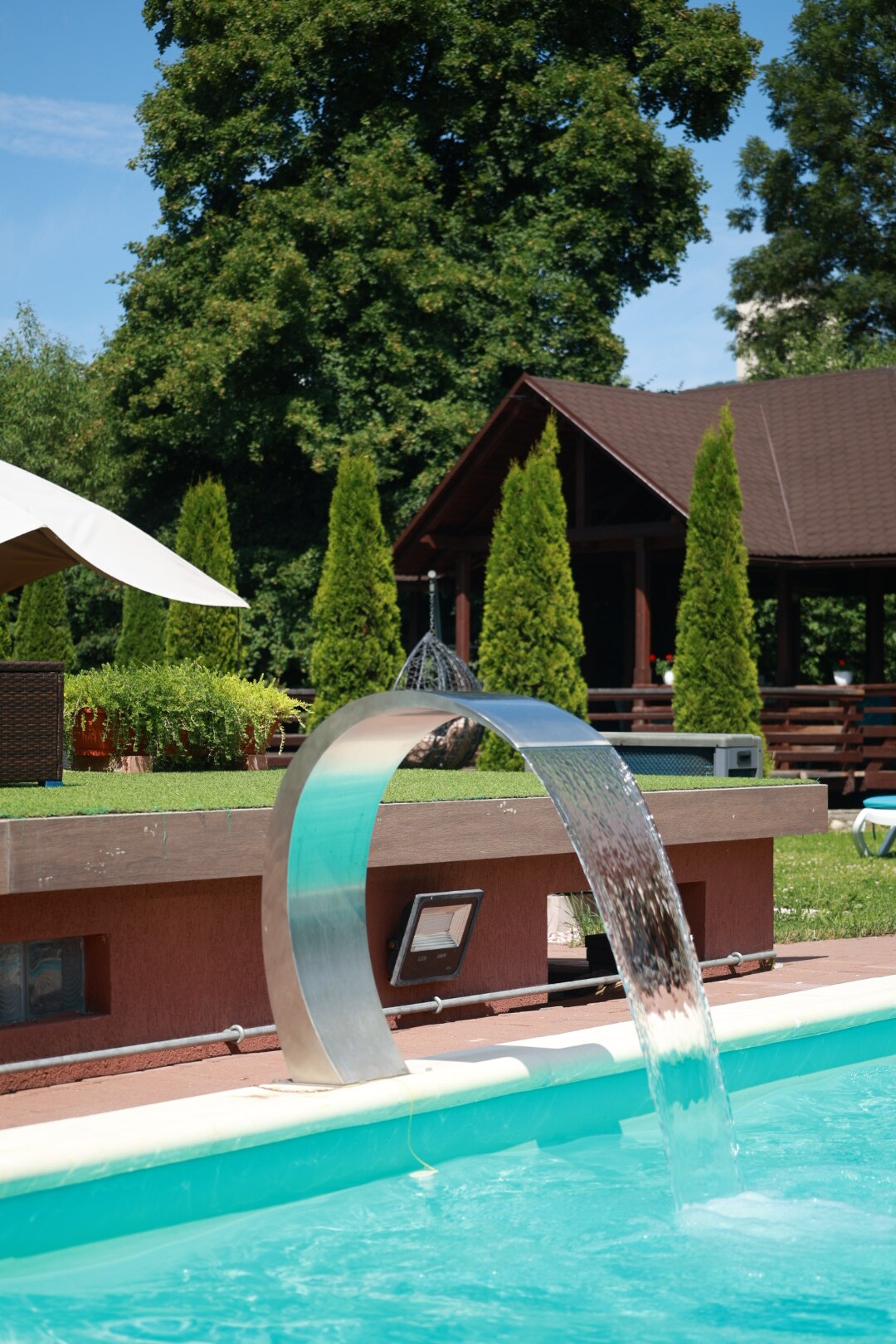 A modern swimming pool features a stainless steel water feature that cascades into the clear water. Surrounding greenery includes well-maintained shrubs and trees. In the background, a rustic wooden structure is partially visible, offering a serene setting.