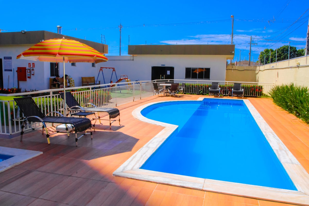 A well-maintained outdoor swimming pool is bordered by a wooden deck. Two lounge chairs and a colorful umbrella are positioned nearby. The surrounding area includes grass and a children's swing set, while clear skies create a bright and inviting atmosphere.