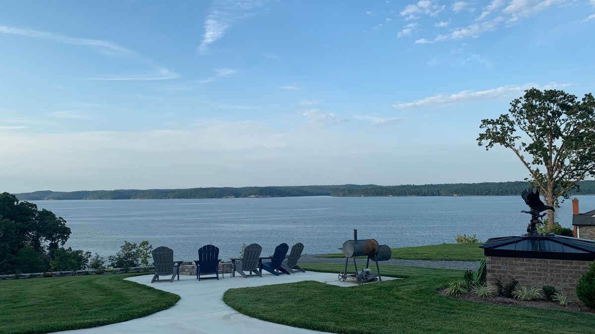 A serene outdoor area is shown, featuring several adirondack chairs arranged near a stone fire pit. The expansive view reveals calm waters and distant hills under a clear blue sky, creating a peaceful setting for relaxation.