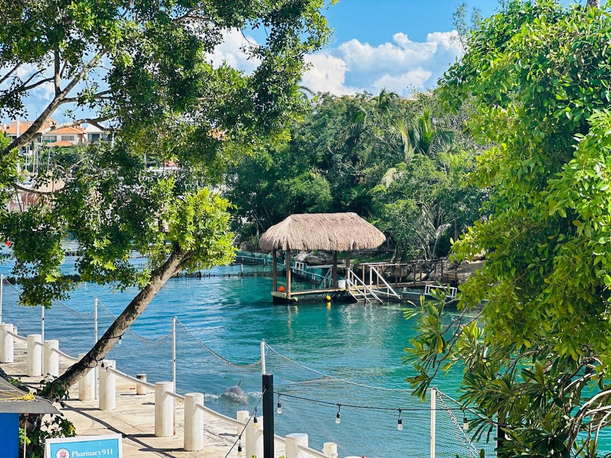 A serene waterfront view showcases a thatched-roof structure on stilts, surrounded by lush greenery. The calm blue water reflects the clear sky, creating a peaceful ambiance. Decorative lights hang along the water's edge, enhancing the inviting outdoor space.