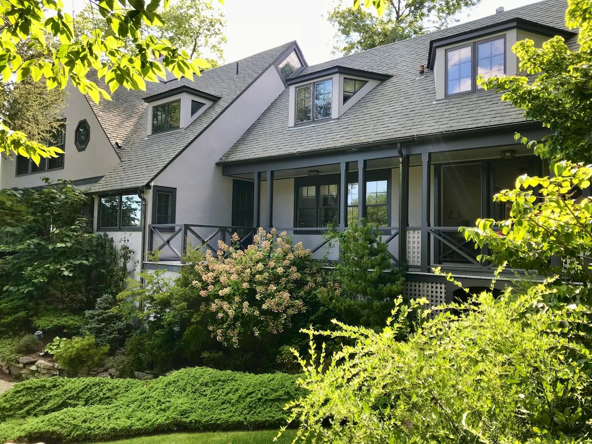 A charming house is nestled among lush greenery, featuring a light-colored exterior with multiple gabled roofs. Large windows allow natural light to brighten the spaces inside. Vibrant flower bushes and neatly trimmed shrubs enhance the home's inviting surroundings.