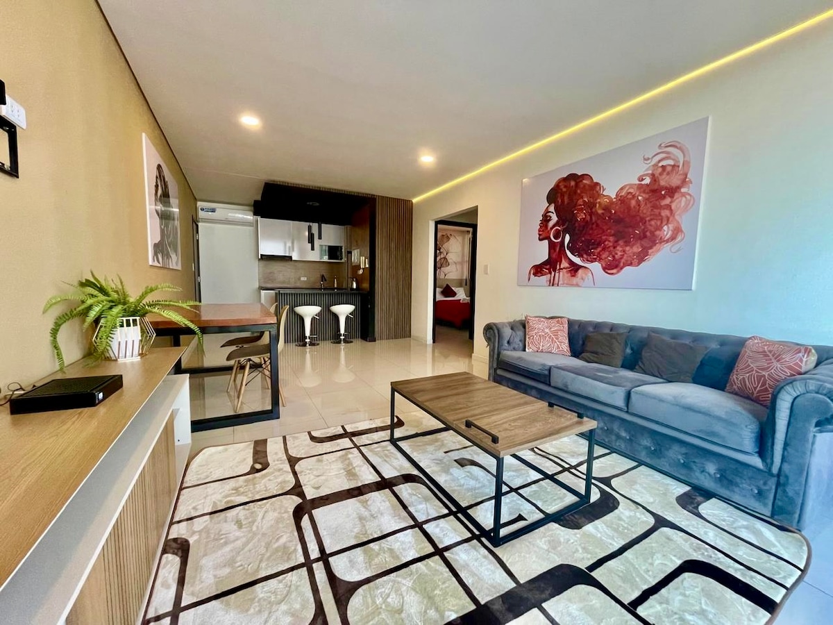 A spacious living area is visible, featuring a comfortable sofa adorned with decorative cushions. A coffee table sits on a patterned rug, while modern bar stools are positioned near a sleek kitchen area in the background. Light fixtures enhance the contemporary decor.