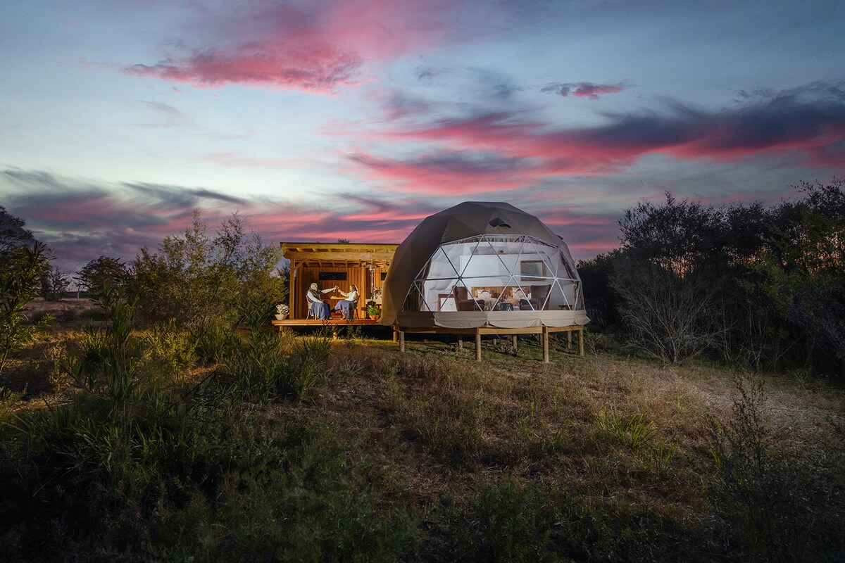 A dome structure is set amidst natural bushland, with a wood deck extending into a tranquil outdoor space. Large windows showcase interior comfort while the sunset paints the sky in soft hues, enhancing the peaceful atmosphere of the retreat.