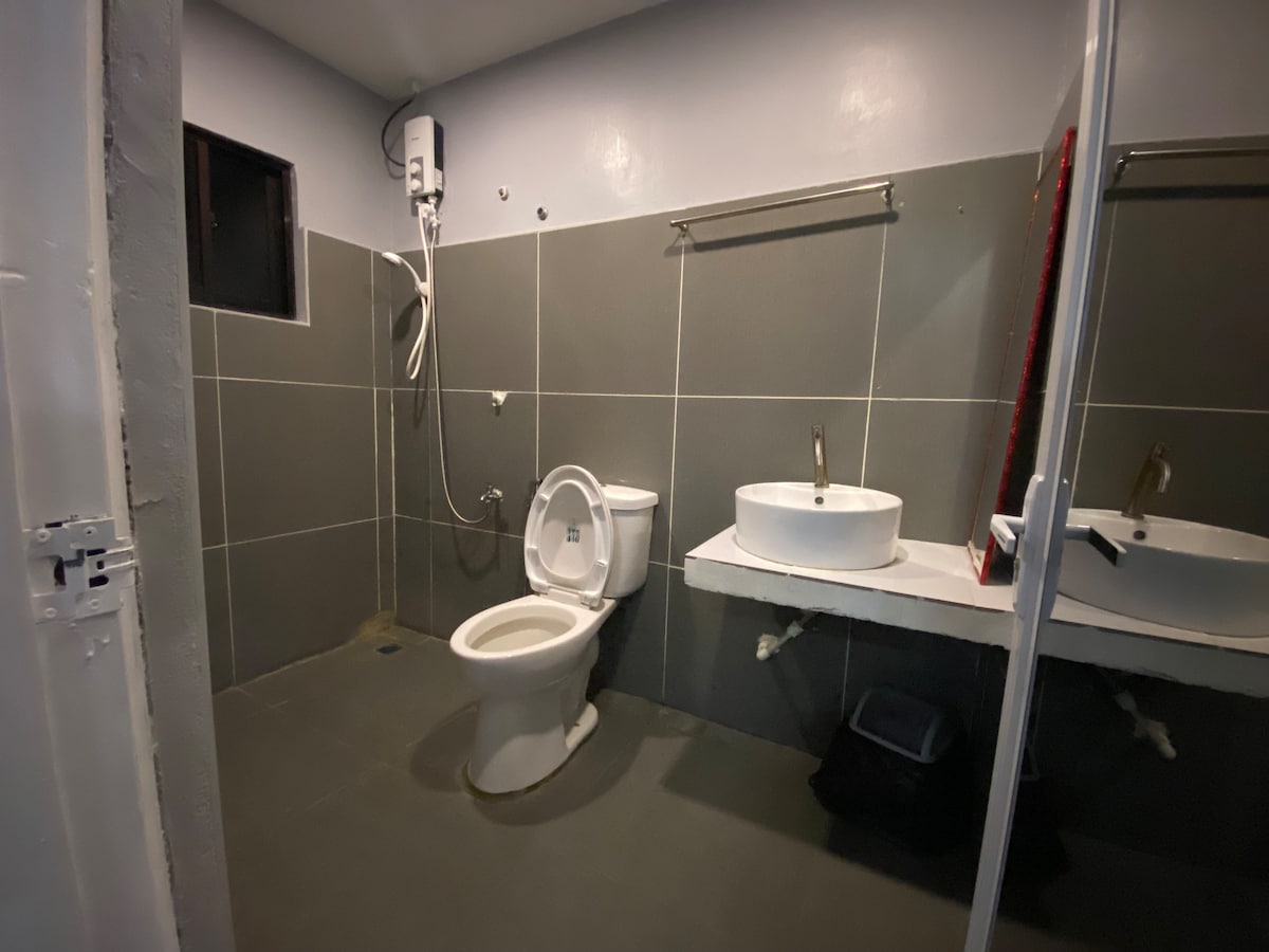A modern bathroom features a walk-in shower, a circular sink atop a countertop, and a toilet. Grey tiles cover the walls and floor, creating a clean and minimalist aesthetic. A small window provides natural light, enhancing the spacious feel of the room.