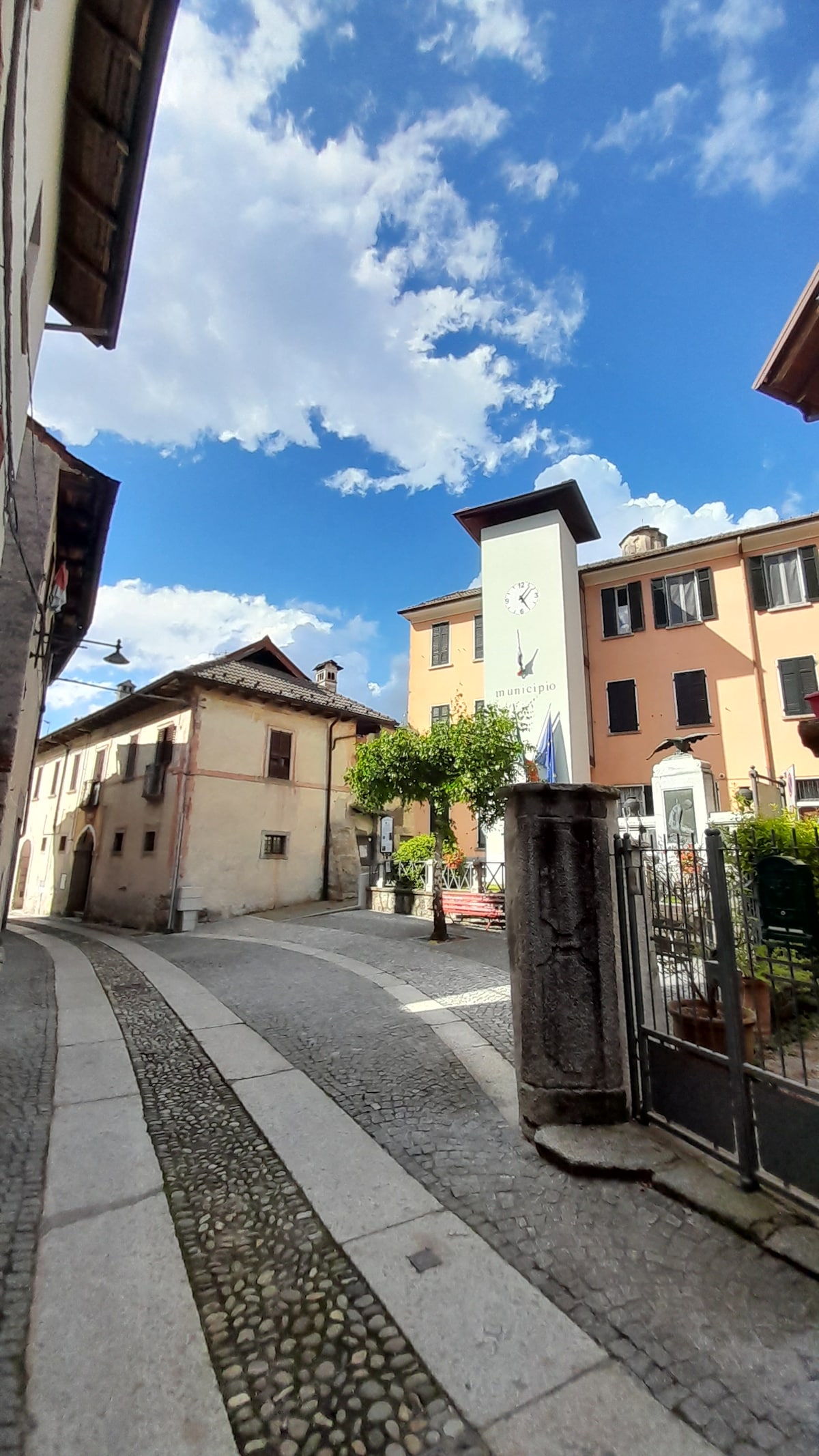 A charming street in a historic area is bordered by traditional buildings featuring varied architecture. A tall, slender structure stands prominently, adorned with graphic elements, while a lush tree adds greenery. The blue sky is punctuated by fluffy white clouds, enhancing the inviting ambiance.