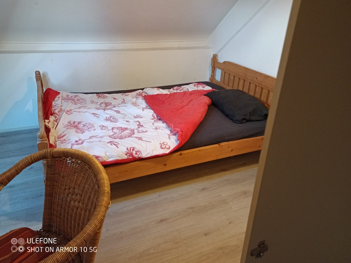 The sleeping area features a wooden bed with a colorful blanket and decorative pillows. A rattan chair is positioned nearby, complementing the natural tones of the wooden floor. Light reflects softly off the sloped ceiling, enhancing the cozy ambiance of the room.
