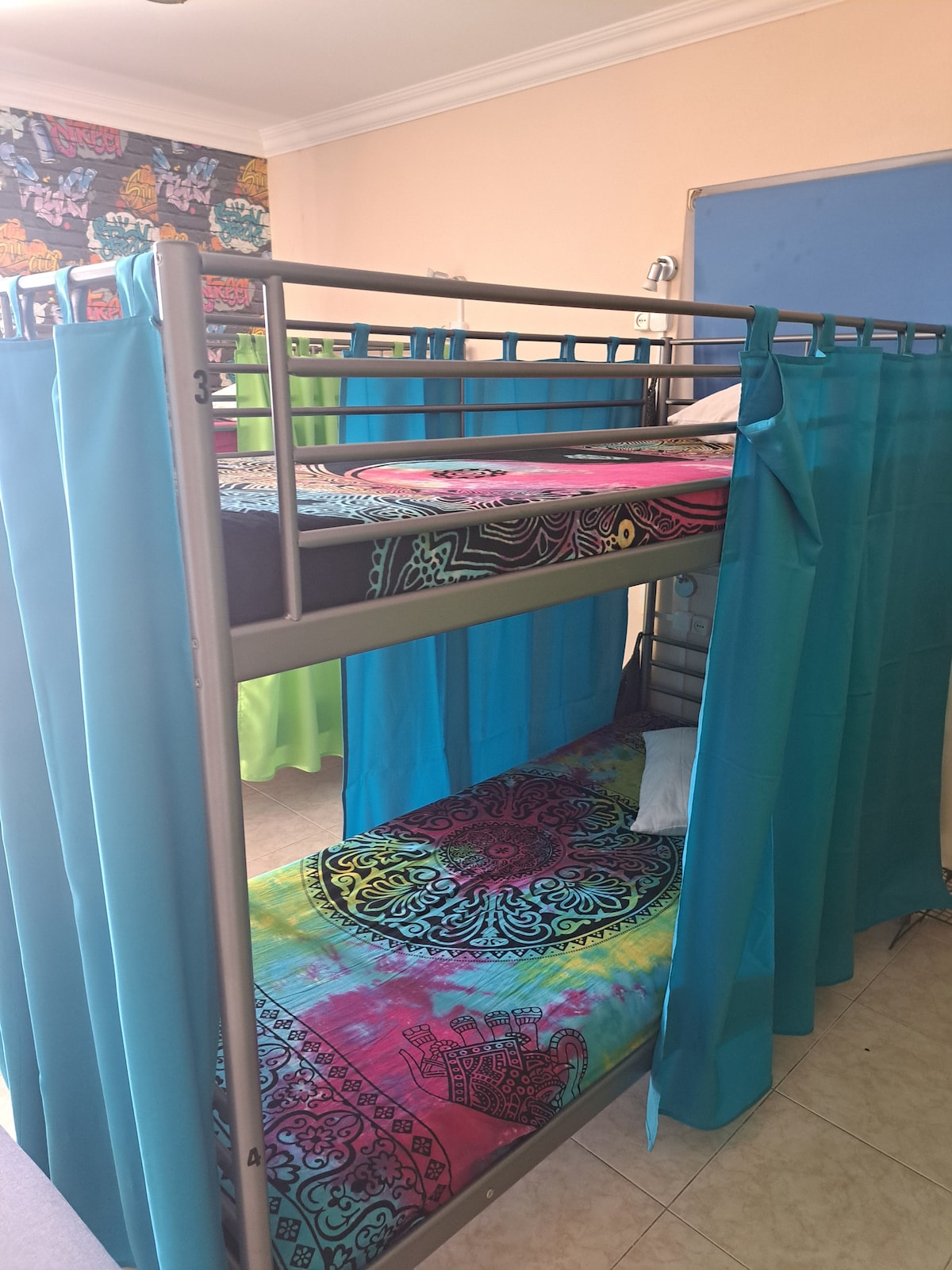 A metal bunk bed is positioned in a brightly colored room. The lower and upper beds are adorned with vibrant, patterned bedding in a tie-dye style. Each bed is enclosed by turquoise curtains, offering privacy and a cozy ambiance.