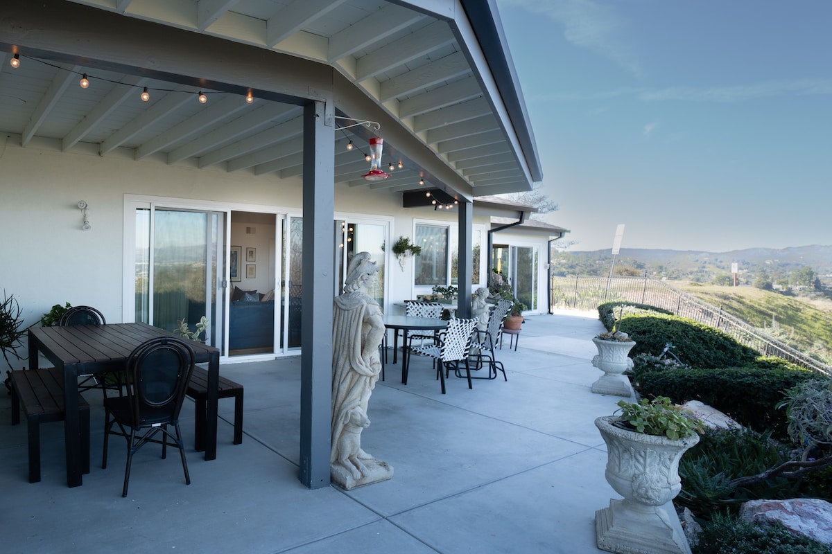 An outdoor patio features a wooden dining table surrounded by black chairs. Potted plants and a decorative statue are present. A backdrop of rolling hills and a clear sky enhances the space, while string lights provide subtle illumination overhead.