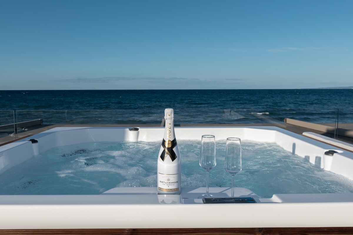 A hot tub is situated on a spacious deck overlooking the sea. A chilled bottle of champagne with two glasses is placed on the edge of the tub. The tranquil waters and clear blue sky create a serene ambiance.