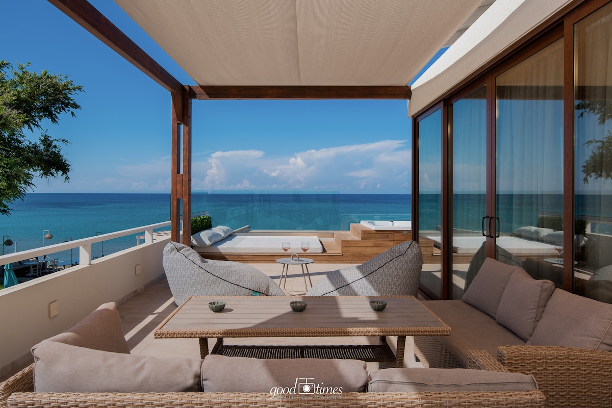 A shaded terrace features a comfortable seating area with modern furniture, offering a stunning view of the sea. The blue hues of the water and sky create a serene backdrop, while a private hot tub is visible on the adjacent deck.