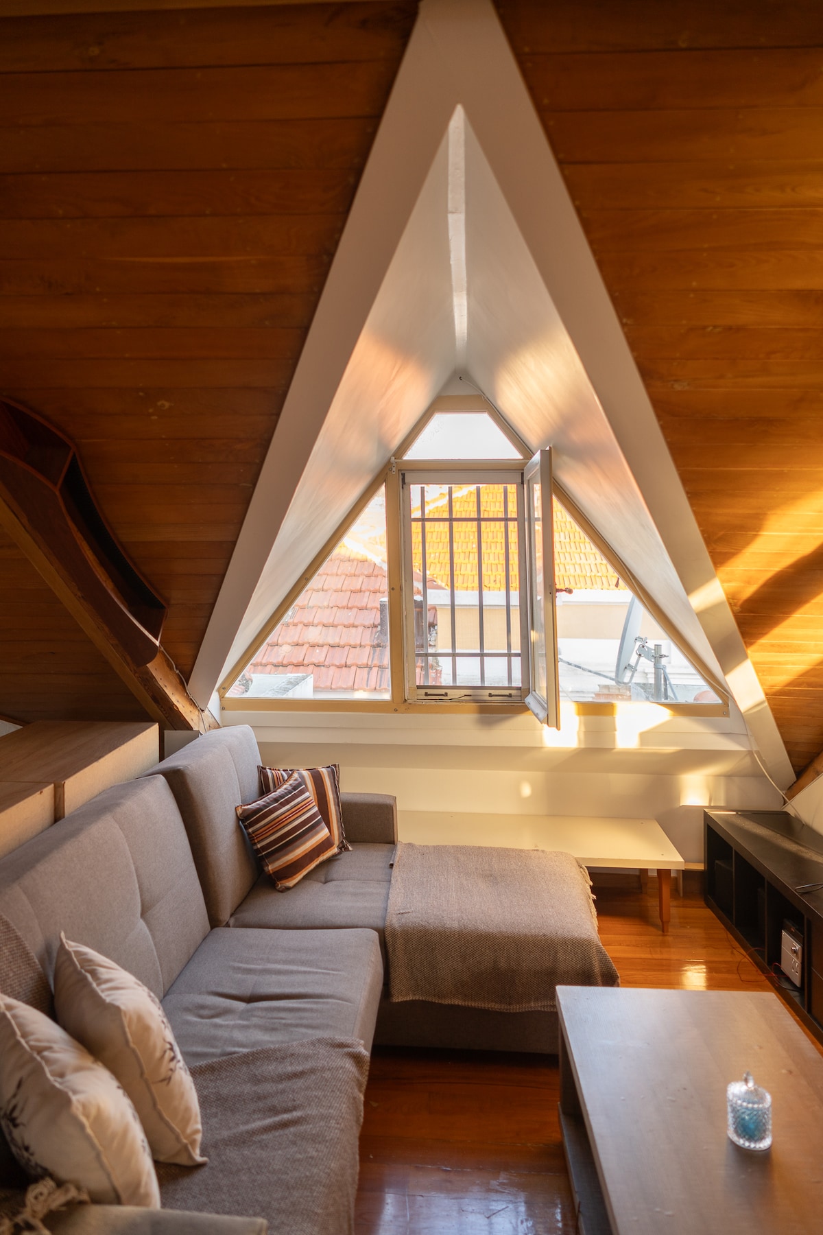 A cozy corner is highlighted by an angular ceiling with wooden beams. A large window allows natural light to fill the space, illuminating a comfortable sofa and simple coffee table. Decorative pillows add subtle texture to the sofa, creating an inviting spot for relaxation.