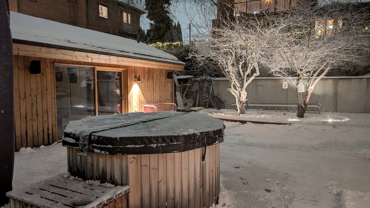 A wooden outdoor hot tub is surrounded by a landscaped area covered in snow, with two lightly frosted trees visible nearby. Soft lighting illuminates the space, highlighting the cozy atmosphere created by the wooden structure in the background.