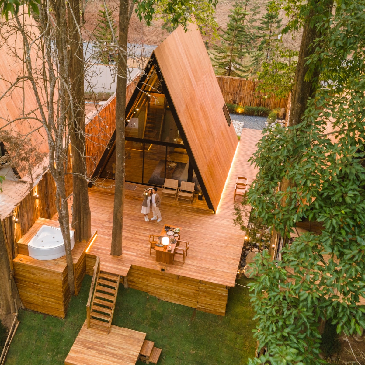 An A-frame wooden cabin is set amidst tall trees, featuring an expansive wooden deck. A hot tub is visible beside a dining area, with two individuals nearby. Soft lighting highlights the structure, creating a seamless connection with the surrounding greenery.