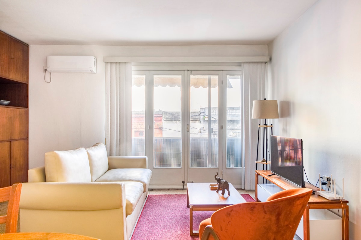 A well-lit living area features a light-colored sofa and wooden seating, complemented by a coffee table and decorative items. Large glass doors open to a balcony, allowing natural light to fill the space, with soft drapes partially drawn.