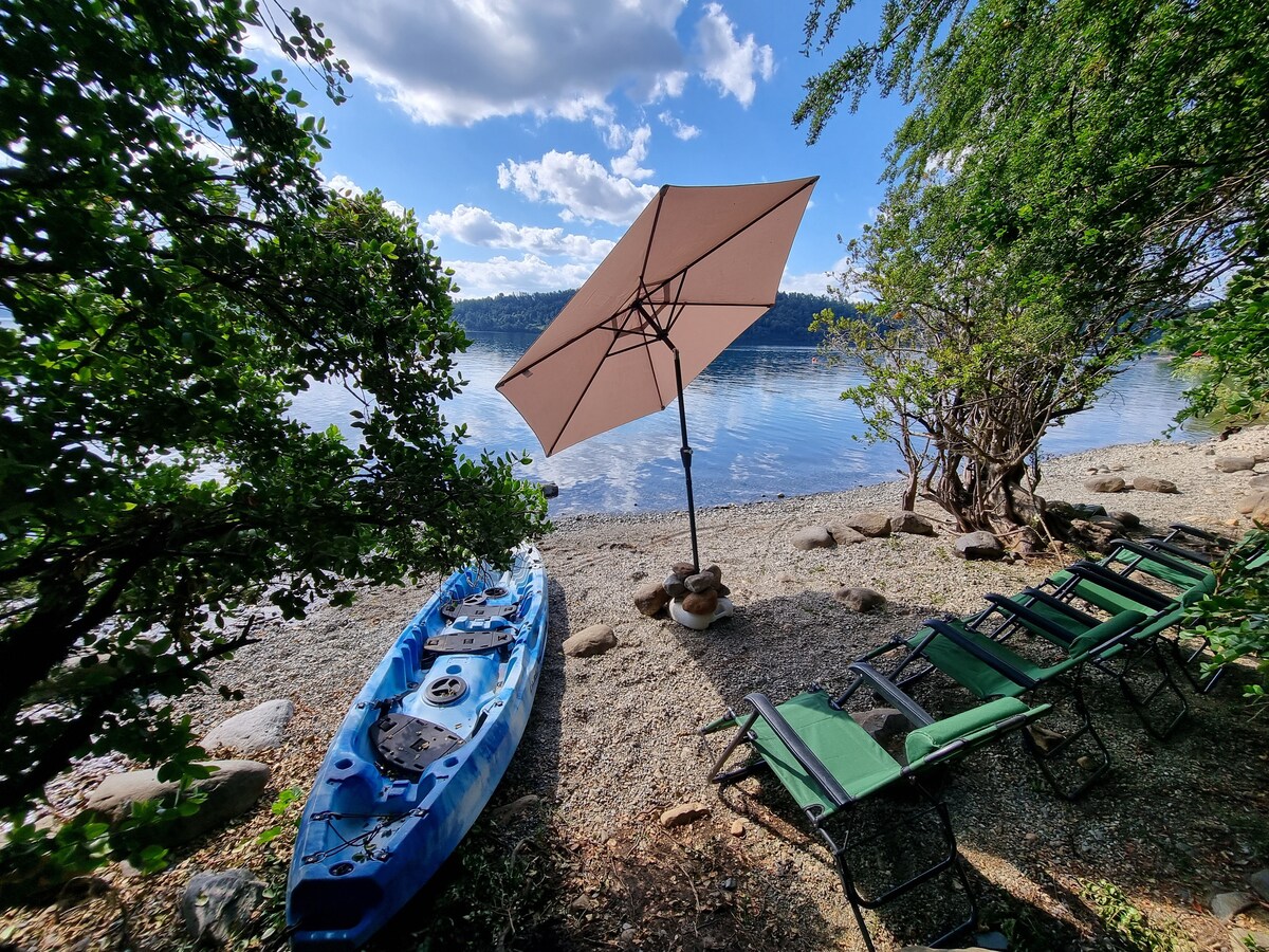 A tranquil lakeside scene is featured with a blue kayak resting on a pebbled beach. A large umbrella provides shade over two green lounge chairs positioned nearby, and trees frame the view of the calm water and blue sky.