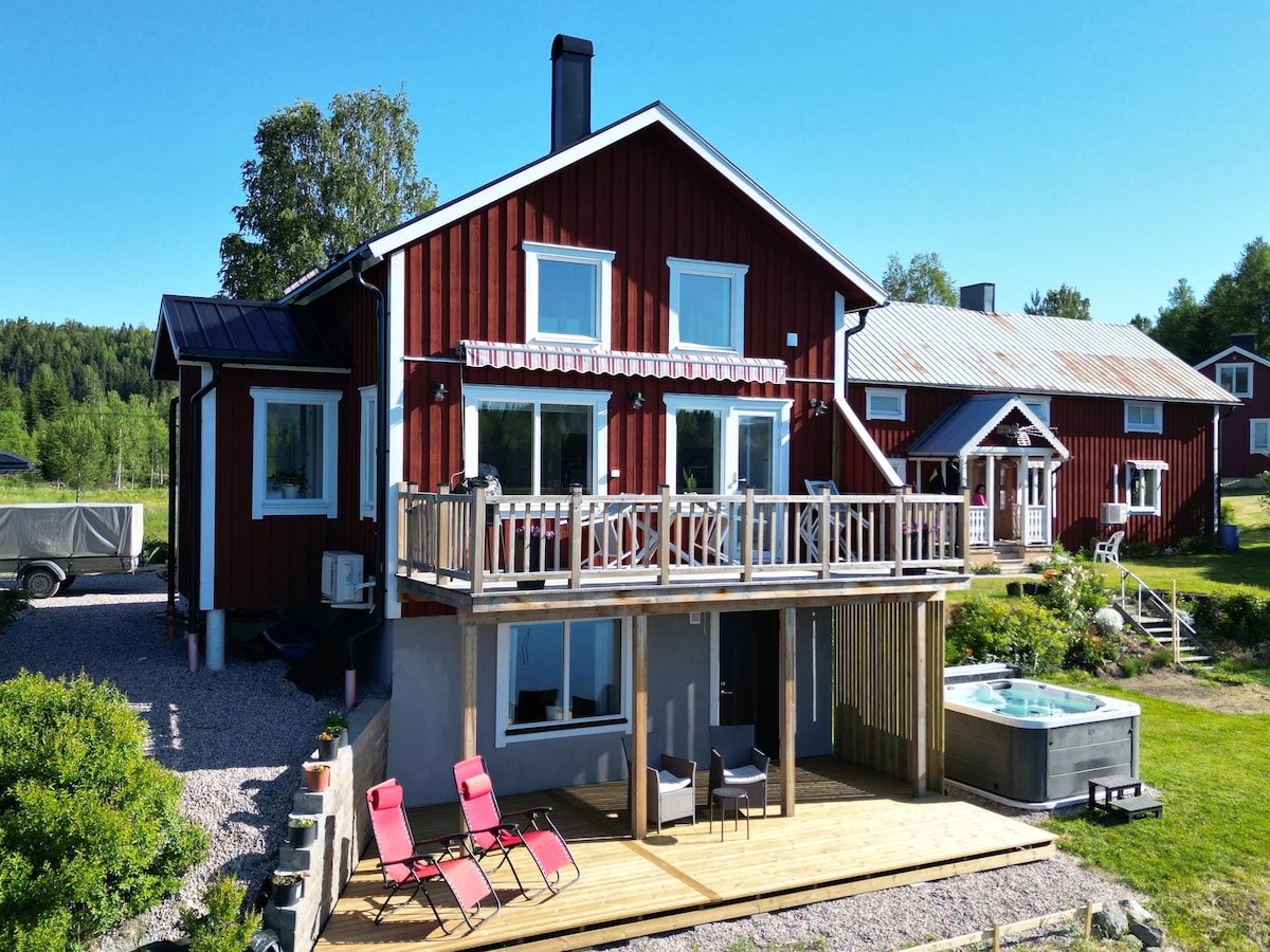 A red wooden house is highlighted by bright blue skies. The front deck features a seating area with two loungers, accompanied by a hot tub to the side. Large windows provide a view of the surrounding greenery, enhancing the outdoor living space.