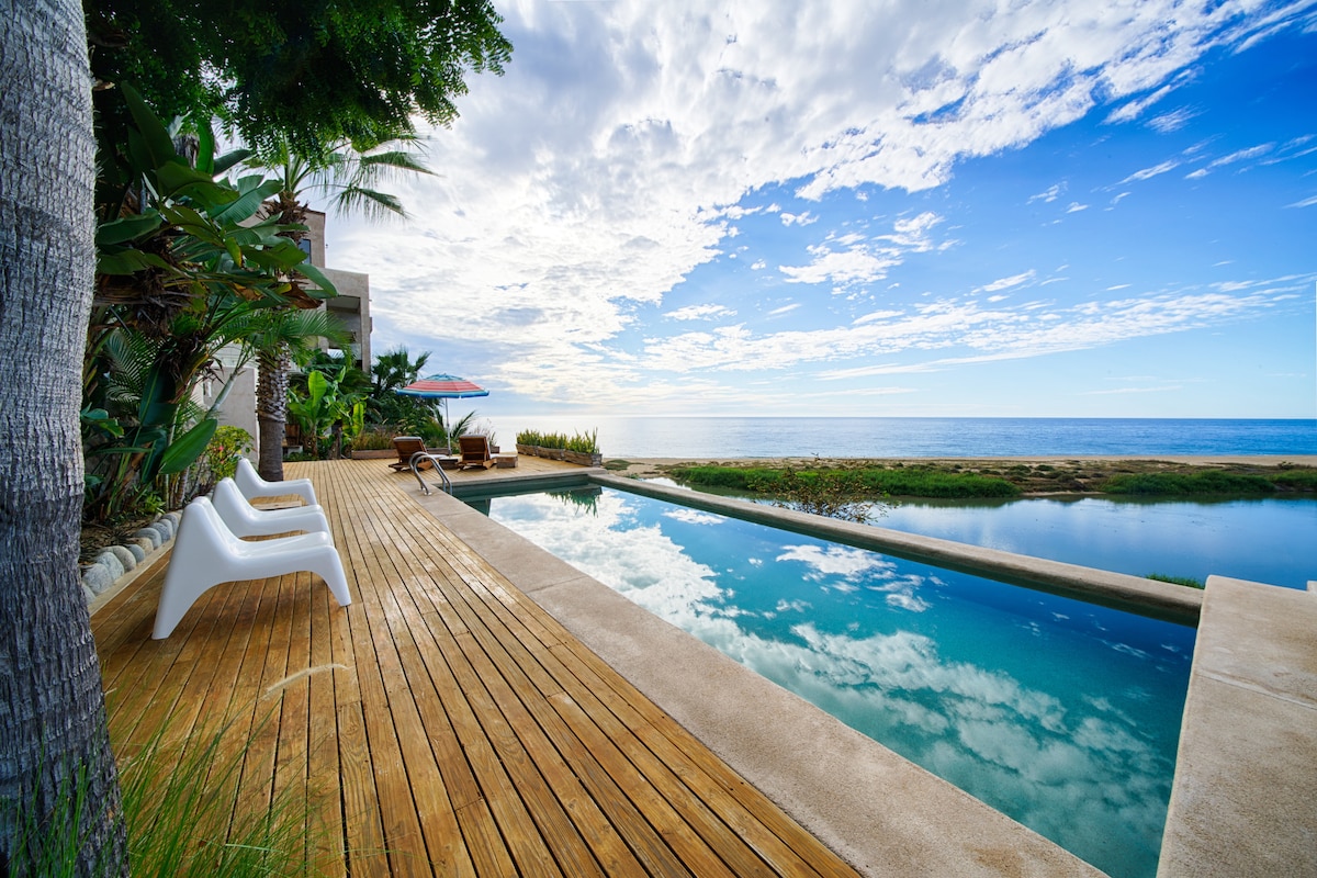 A clear swimming pool stretches across the deck, reflecting the open sky. Comfortable sun loungers are positioned nearby, alongside vibrant greenery. The tranquil ocean is visible in the background, complementing the serene atmosphere created by the expansive wooden patio.