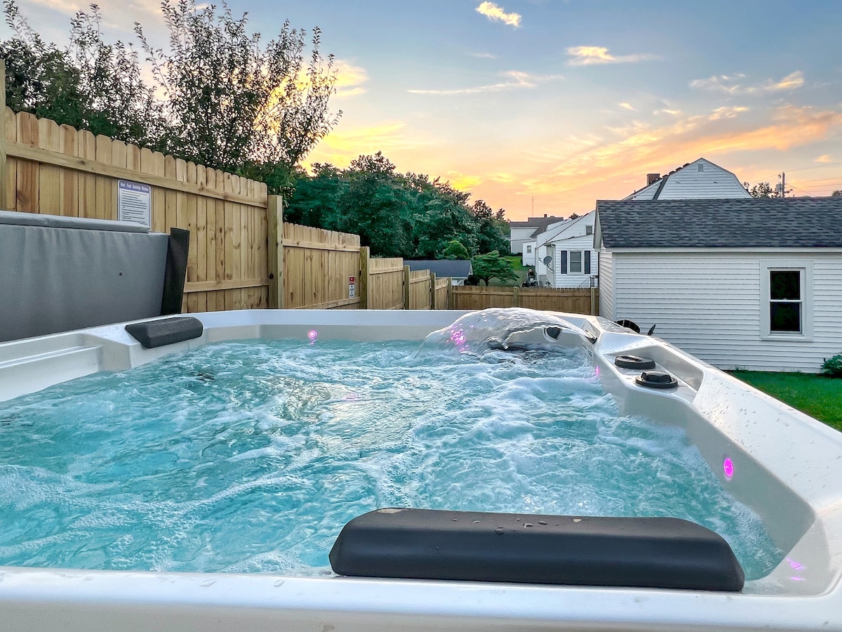 A bubbling hot tub is positioned in the backyard, with clear water reflecting the sunset's colors. Surrounding wooden fencing creates a sense of privacy, while a gentle glow from the spa's lights enhances the serene, relaxing atmosphere.