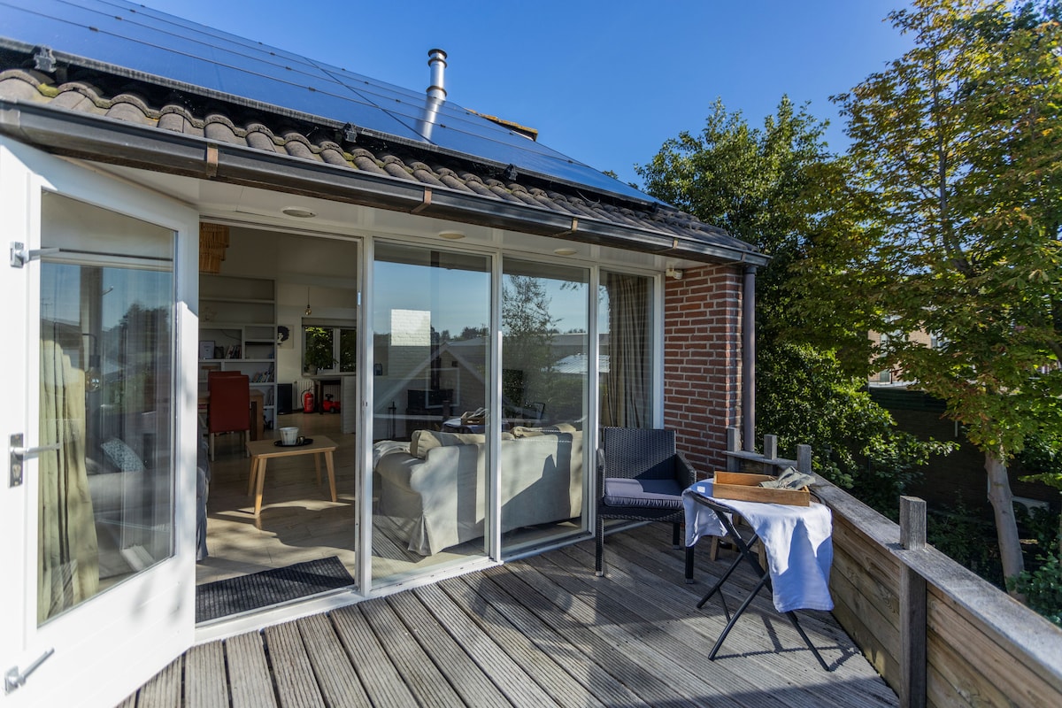 A sunny terrace is featured, extending from the living area with glass doors providing natural light. Outdoor furniture includes a small table and two chairs, set against a backdrop of greenery. Solar panels are visible on the roof, reflecting a commitment to sustainability.