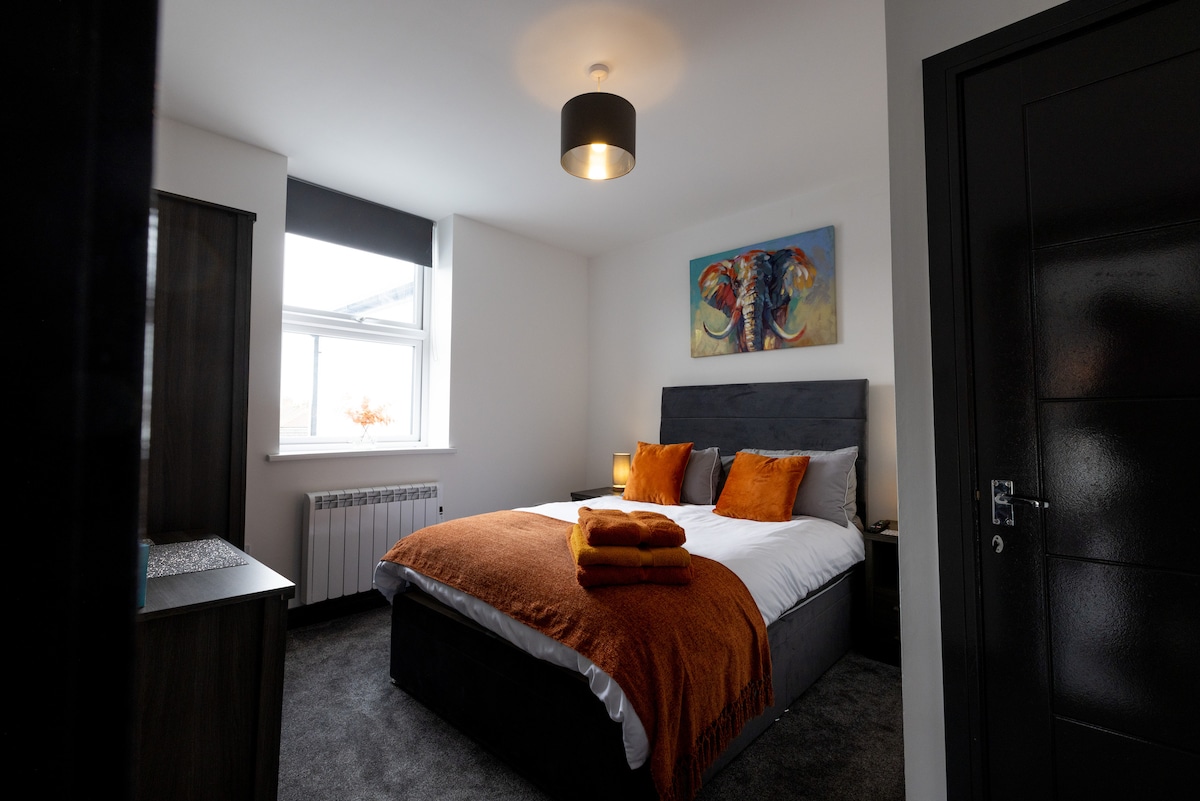 A well-appointed bedroom features a spacious bed layered with crisp white linens and orange accent pillows. An orange throw is neatly folded at the foot of the bed. A window allows natural light to fill the room, complemented by contemporary artwork on the wall.