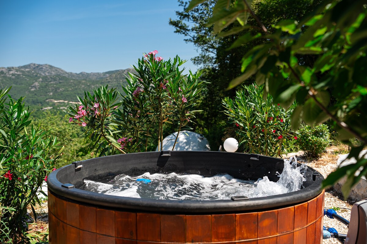 A wooden hot tub is filled with bubbling water, surrounded by lush green plants and blooming flowers. Scenic mountain views provide a serene backdrop, enhancing the outdoor relaxation space.
