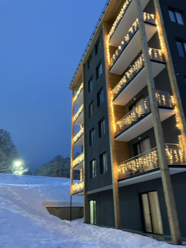 A modern building is depicted in the evening hours, illuminated by warm white lights that outline the balconies. Snow blankets the ground, complementing the surrounding winter landscape. Soft lighting from the building enhances the tranquil atmosphere of the scene.