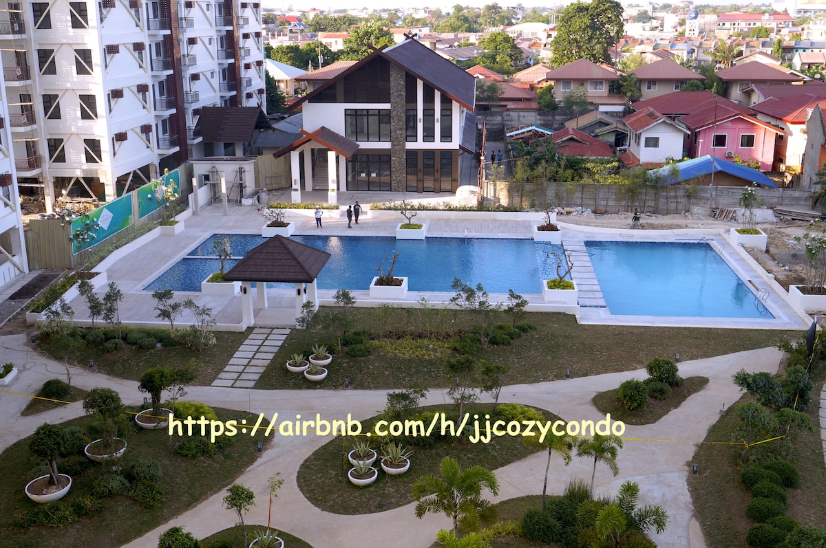 The image showcases a well-maintained outdoor area featuring two swimming pools, surrounded by neatly landscaped gardens with paths and planters. A modern building structure is visible in the background, along with residential houses beyond the property, contributing to a serene environment.