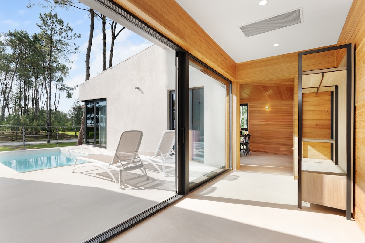 Large glass doors open to a serene outdoor space featuring a private pool. Sun loungers are positioned nearby, surrounded by a wooden terrace and glimpses of pine trees. The interior showcases wooden paneling and a well-lit area, enhancing the connection between indoor and outdoor settings.