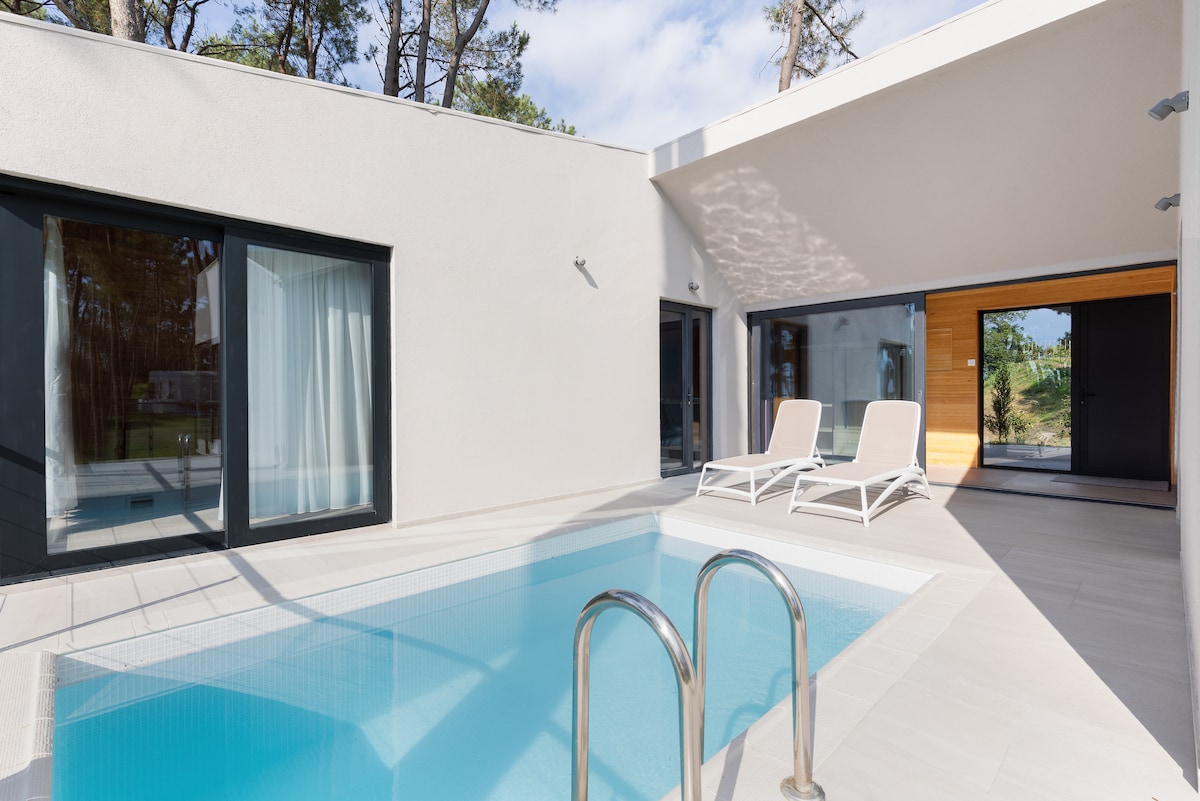 A private pool is framed by modern architecture, featuring sleek white walls and large glass doors. Sun loungers are positioned nearby, providing a space for relaxation. Surrounding greenery is visible through the windows, enhancing the tranquil atmosphere.