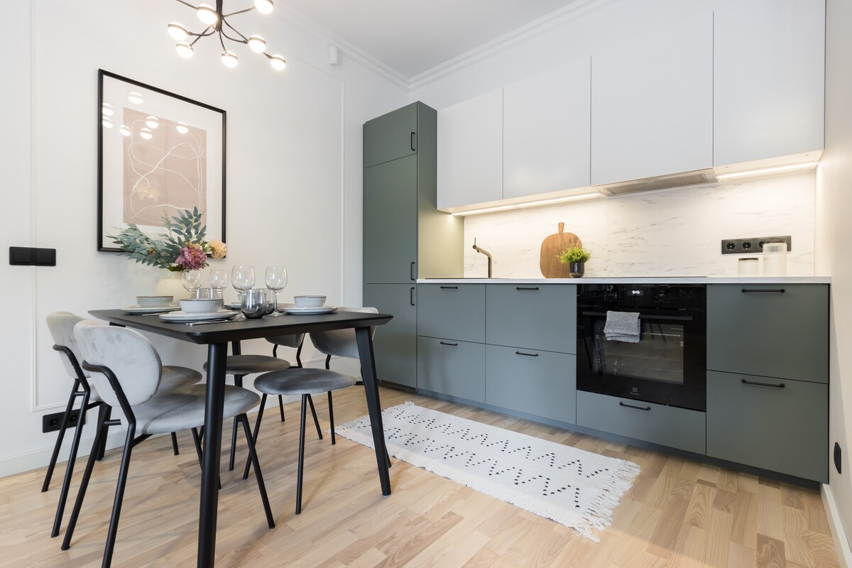 A modern kitchen is presented with sleek cabinetry in muted tones. A dining table is set for four with elegant dishware. Warm lighting illuminates the space, highlighting the marble backsplash and contemporary appliances, creating a functional and inviting environment.