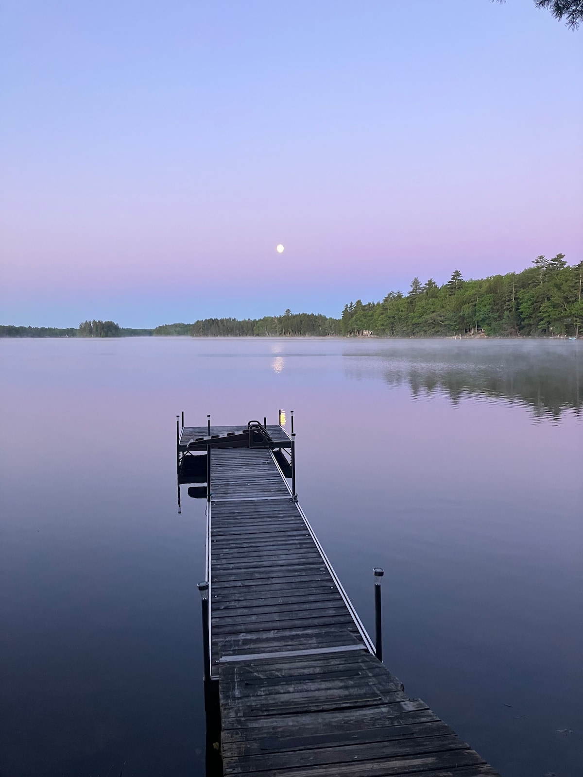 A wooden dock extends into a calm lake, reflecting pastel hues of dawn. The full moon is visible above, surrounded by a mist that hovers over the water's surface. Lush greenery lines the opposite shore, enhancing the tranquil scene.