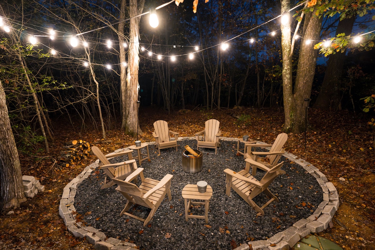 An outdoor gathering area is illuminated by string lights, surrounding a circular fire pit. Multiple wooden Adirondack chairs are arranged on gravel, inviting relaxation in a wooded setting. The ground is adorned with fallen leaves, enhancing the natural ambiance.