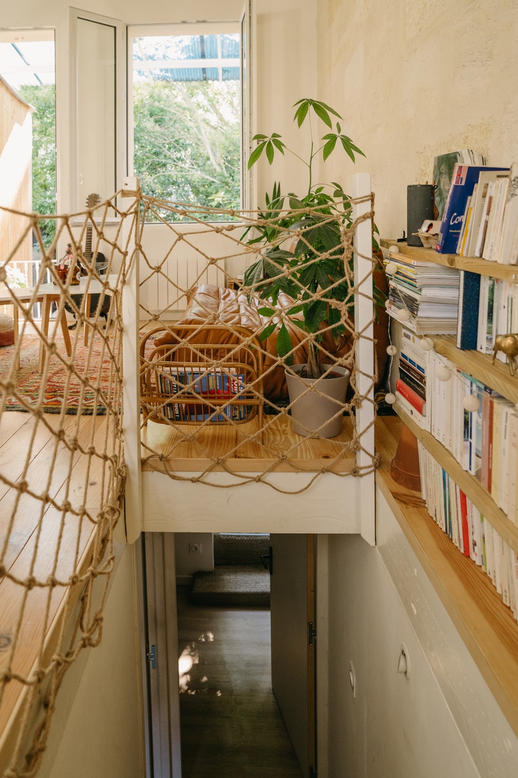 An interior space showcases a suspended walkway adorned with a netting feature, connecting different levels. A variety of books line wooden shelves on one side, while greenery adds a natural touch. Soft light filters through open doors, enhancing the sense of openness.
