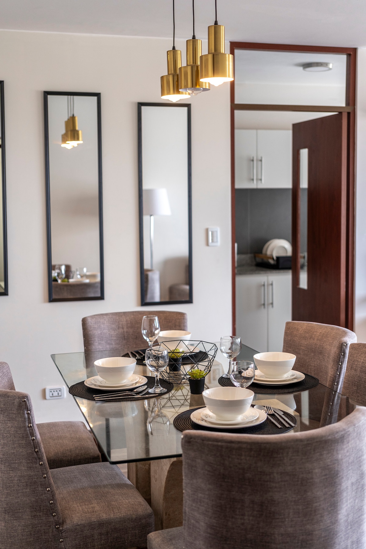 A dining area features a glass table surrounded by upholstered chairs. A simple table setting includes plates, bowls, and glasses, complemented by a small decorative plant in the center. The room is illuminated by pendant lights and mirrors reflect the bright space.